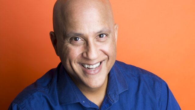 Smiling man in blue shirt, orange background.